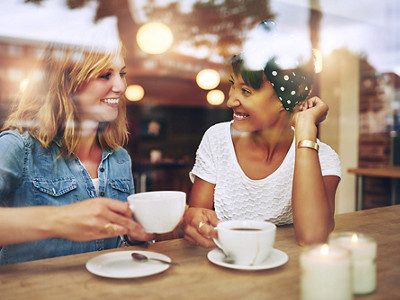 Two multi ethnic friends enjoying coffee together in a coffee shop viewed through glass with reflections as they sit at a table chatting and laughing