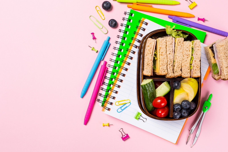 Flat lay with wooden lunch box with sandwiches, vegetables and fruits on pink background and school supplies. Healthy children eating concept. Top view with copy space.